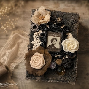 May include: A rectangular book with a dark, textured cover decorated with fabric flowers in cream and brown, a black ornate frame containing a vintage portrait of a person, white lace trim, and various dark buttons and circular embellishments.