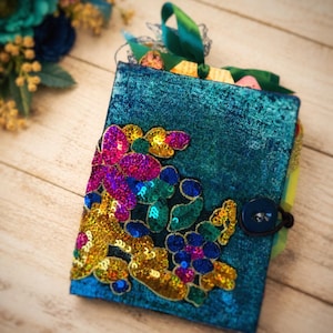 May include: A rectangular journal with a textured teal cover featuring an appliqué of vibrant, multi-colored sequins in shades of gold, pink, blue, and green. The journal is secured with a dark blue button and an elastic loop, with green ribbons.