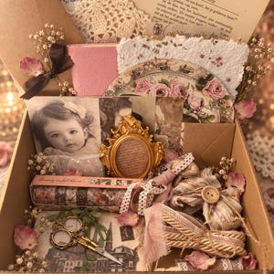 May include: A cardboard gift box containing a collection of vintage-style items including a black and white photograph of a child, a gold ornate frame, a pair of gold scissors, rolls of floral washi tape, lace ribbons, dried flowers, and paper ephemera.