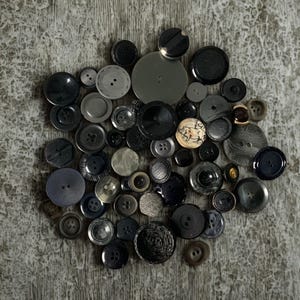 以下が含まれることがあります： さまざまなサイズと質感の黒とグレーのボタンのコレクション。ボタンはクラスター状に配置され、平らな円盤からドーム型のデザインまで、さまざまな形状を示しています。一部のボタンには、縫い穴が見えます。