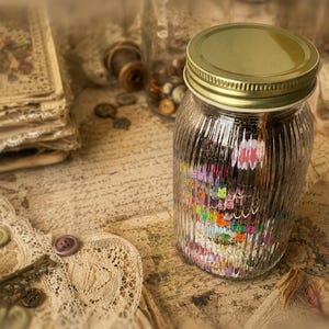 May include: A clear, vertically ribbed glass jar with a gold-colored metal lid. The jar is filled with a collection of small, multi-colored craft beads. The glass has a textured, fluted surface that distorts the view of the colorful contents inside.