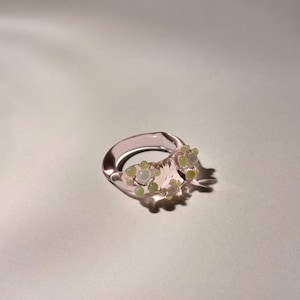 May include: A transparent pink ring with a floral design. The ring features small, round, light green and white embellishments clustered on the band. The ring is set against a soft, off-white background, creating a delicate and feminine aesthetic.