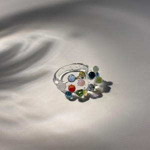 以下が含まれることがあります： カラフルな丸いビーズで飾られた透明なガラスリング。ビーズは青、緑、赤、ピンク、黄色の色合いです。リングは影のある白い背景に配置されています。