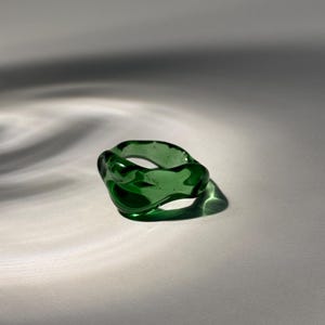 Könnte beinhalten: Ein transparenter, smaragdgrüner Ring mit einer organischen, unregelmäßigen Form. Der Ring besteht aus Glas und wirft einen Schatten auf eine weiße Oberfläche. Das Design ist modern und minimalistisch.