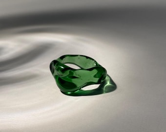 May include: A transparent, emerald green ring with an organic, irregular shape. The ring is made of glass and casts a shadow on a white surface. The design is modern and minimalist, suitable for everyday wear.