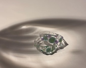 Op de afbeelding: Een transparante glazen ring met groene en paarse accenten. De ring is gemaakt van glas en heeft een unieke, organische vorm.