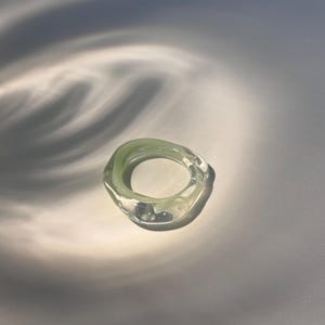 May include: A clear, pale green ring with an organic, irregular shape. The ring appears to be made of glass or a similar transparent material. The ring is set against a white background with soft shadows.