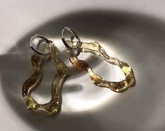May include: A pair of gold and clear resin hoop earrings with a wavy, organic shape. The earrings are made of a translucent material with a subtle shimmer.