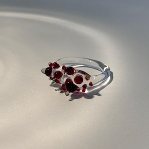 Puede incluir: Un anillo de vidrio transparente con detalles rojos y verdes. El anillo presenta pequeñas cuentas redondas de vidrio rojo y algunas cuentas verdes. El anillo está sobre un fondo blanco, con sombras que añaden profundidad.
