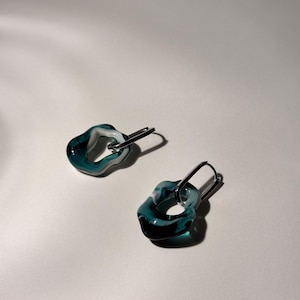Op de afbeelding: Een paar oorbellen van teal en wit gemarmerd glas. Elke oorbel heeft een unieke, organische vorm met een zilverkleurige metalen sluiting. De oorbellen worden gepresenteerd op een witte ondergrond.