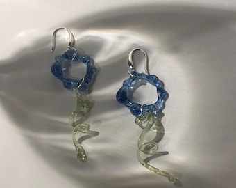 May include: A pair of drop earrings featuring a blue glass ring with a textured, organic edge. Attached to each blue ring is a pale green, translucent glass spiral that hangs downward. The earrings are suspended from silver-toned metal hooks.