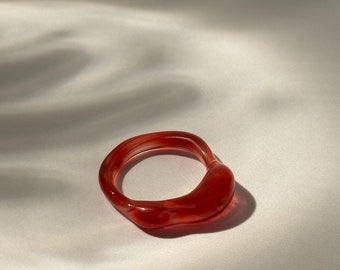 Può includere: Un anello di vetro rosso con una forma unica e irregolare. L'anello è liscio e lucido, con una leggera traslucenza.