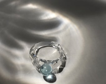 Può includere: Un anello di vetro trasparente con un design testurizzato e una perla di vetro blu chiaro attaccata alla parte anteriore.
