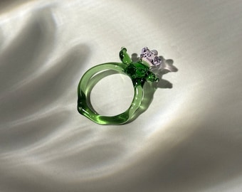 May include: A green glass ring with a purple flower on top. The ring is handmade and has a unique, organic shape.