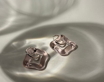 May include: A pair of pink glass hoop earrings with a square shape. The earrings are made of a translucent glass with a slightly cloudy appearance.