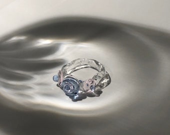 May include: A clear glass ring with pink and blue glass flowers attached to it. The ring is sitting on a white surface.