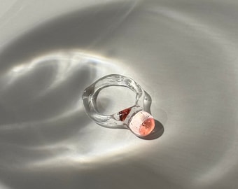 Può includere: Un anello di vetro trasparente con un accento di vetro rosa. L'anello è su una superficie bianca.