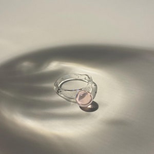 May include: A clear glass ring with a pink glass orb attached. The ring is made of clear glass with small air bubbles throughout. The orb is a light pink color and is smooth.