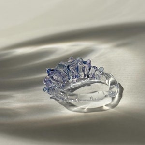 以下が含まれることがあります： 白の表面に置かれた、テクスチャ加工された表面と紫と青のデザインが施された透明なガラスのリングです。