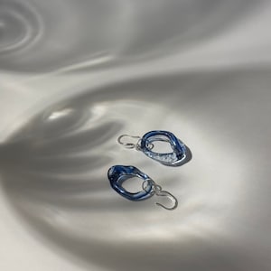 Peut inclure: Une paire de boucles d'oreilles en verre bleu avec une forme irrégulière unique. Les boucles d'oreilles sont suspendues à des crochets argentés.