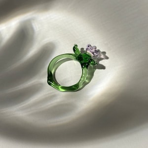 Op de afbeelding: Een groene glazen ring met een paarse bloem erop. De ring is handgemaakt en heeft een unieke, organische vorm.