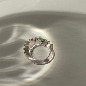 May include: A clear glass ring with green accents. The ring is thick and has a rounded shape.