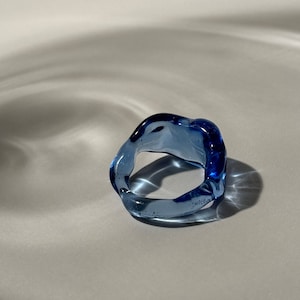 Puede incluir: Un anillo de vidrio azul con una forma irregular y orgánica. El anillo es translúcido y tiene un ligero brillo.