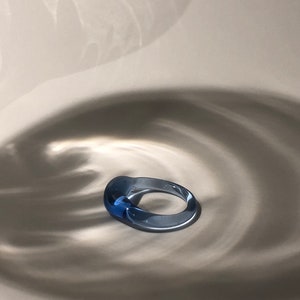 May include: A transparent blue ring with a rounded, oval shape. The ring is made of glass or a similar material, and it sits on a light-colored surface. The lighting creates shadows that add depth and dimension to the image.