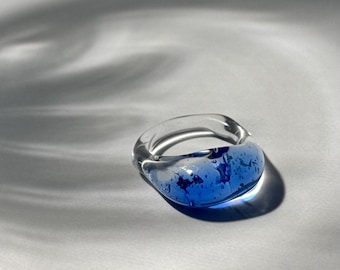 May include: A clear glass ring with a rounded band and a blue, speckled design. The ring is set against a white background with soft shadows, highlighting its transparent and colorful features. The ring is a fashion accessory.