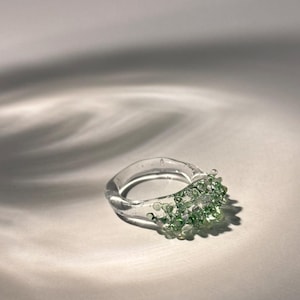 May include: A clear glass ring with a band of small, light green beads clustered on the top. The ring is set against a white background with shadows.
