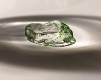 May include: A clear green glass ring with air bubbles trapped inside. The ring is irregularly shaped and has a wavy, organic design.