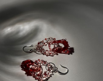May include: A pair of red glass earrings with a teardrop shape and a silver hook. The earrings are made of small glass beads that are strung together to form a delicate design.
