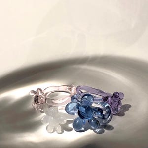 Op de afbeelding: Drie glazen ringen met een bloemvormig ontwerp. De ringen zijn in tinten roze, paars en blauw.