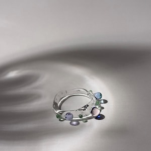 Op de afbeelding: Een heldere glazen ring met kleurrijke, bolvormige versieringen. De ring heeft blauwe, groene en paarse glazen accenten, wat een speels ontwerp creëert. De ring staat tegen een neutrale achtergrond.