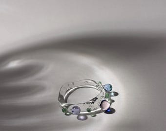 Può includere: Un anello di vetro trasparente adornato con abbellimenti sferici colorati. L'anello presenta accenti in vetro blu, verde e viola, creando un design giocoso. L'anello è posto su uno sfondo neutro.