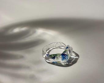 Op de afbeelding: Een transparante glazen ring met blauwe en groene swirls erin. De ring ligt op een witte achtergrond.