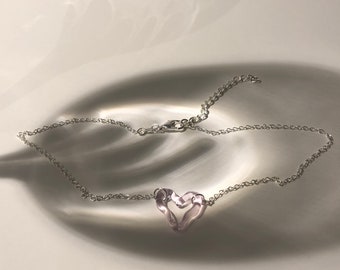 May include: A delicate silver chain necklace with a heart-shaped pendant. The pendant is a soft pink color and appears to be made of glass or a similar translucent material. The necklace is displayed on a reflective surface.