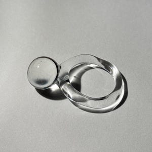 Puede incluir: Un anillo de vidrio transparente que presenta una esfera lisa unida a una banda de forma orgánica ligeramente irregular. El material translúcido refleja la luz, creando sombras y reflejos sutiles en las superficies curvas del objeto.
