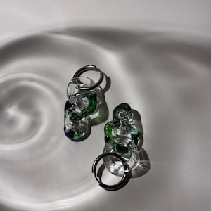 May include: A pair of silver hoop earrings with green and clear glass chain links. The earrings are on a white surface.