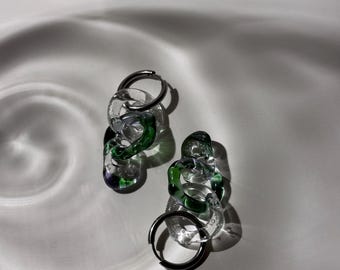 May include: A pair of silver hoop earrings with green and clear glass chain links. The earrings are on a white surface.