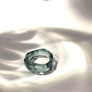 May include: A teal blue glass ring with an irregular, organic shape. The ring is made of clear glass with a slight blue tint and has a rough, textured surface.