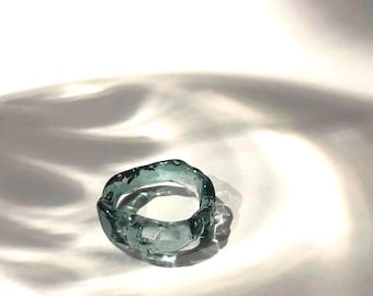 May include: A teal blue glass ring with an irregular, organic shape. The ring is made of clear glass with a slight blue tint and has a rough, textured surface.