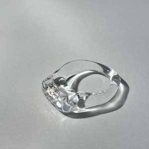 May include: A clear, transparent ring with an irregular, organic shape. The band features a smooth, glossy finish that catches the light, creating subtle prismatic reflections within the material. The design is solid and continuous throughout.