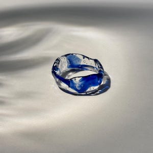 May include: A clear glass ring featuring an irregular, wavy band design with swirling deep blue pigment throughout the material and small air bubbles trapped within the structure, creating a translucent effect with a smooth, polished finish.