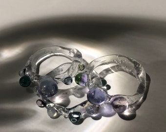 Può includere: Tre anelli di vetro trasparente con perline di vetro viola, blu e verde. Gli anelli sono impilati uno sopra l'altro.