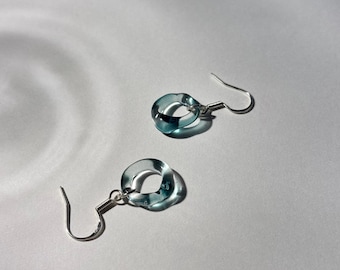May include: A pair of dangle earrings featuring translucent, light blue glass rings suspended from silver-toned metal fishhook wires. The circular glass components have a smooth, rounded profile and a clear, reflective quality that catches light.
