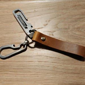 Könnte beinhalten: Ein silberner Multitool-Schlüsselanhänger mit einem Flaschenöffner, einem kleinen Schraubendreher und einem Schlüsselring. Der Schlüsselanhänger ist an einem braunen Lederband mit einem Metallknopf befestigt.