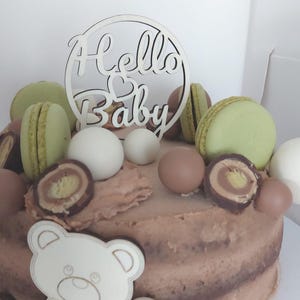 May include: A chocolate frosted cake decorated with green macarons, chocolate candies, and white spheres. A wooden cutout with the words "Hello Baby" and a teddy bear are on the cake. Perfect for a baby shower.