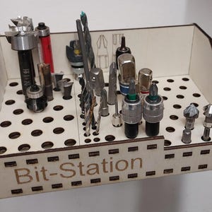 Peut inclure: Un organisateur de rangement pour mèches en bois avec le texte "Bit-Station" sur le devant. L'organisateur est doté d'une grille de trous pour contenir diverses mèches de perçage, mèches de toupie et autres outils.
