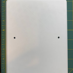 May include: A white rectangular sign with rounded corners and two pre-drilled holes. The sign is made of a smooth, solid material and sits on a green cutting mat with grid lines. The sign is approximately 8 inches tall and 6 inches wide.